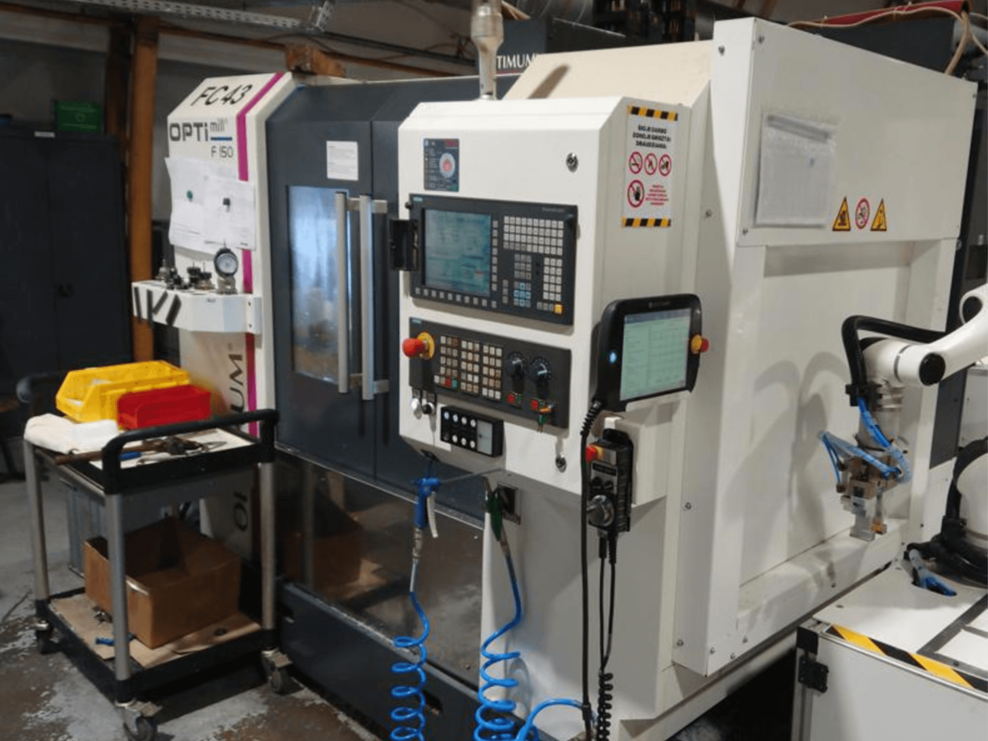 Bedienfeld der OPTIMUM F50 Fräsmaschine mit Schläuchen, Werkzeugen und einem Roboterarm in einer Werkstattumgebung.