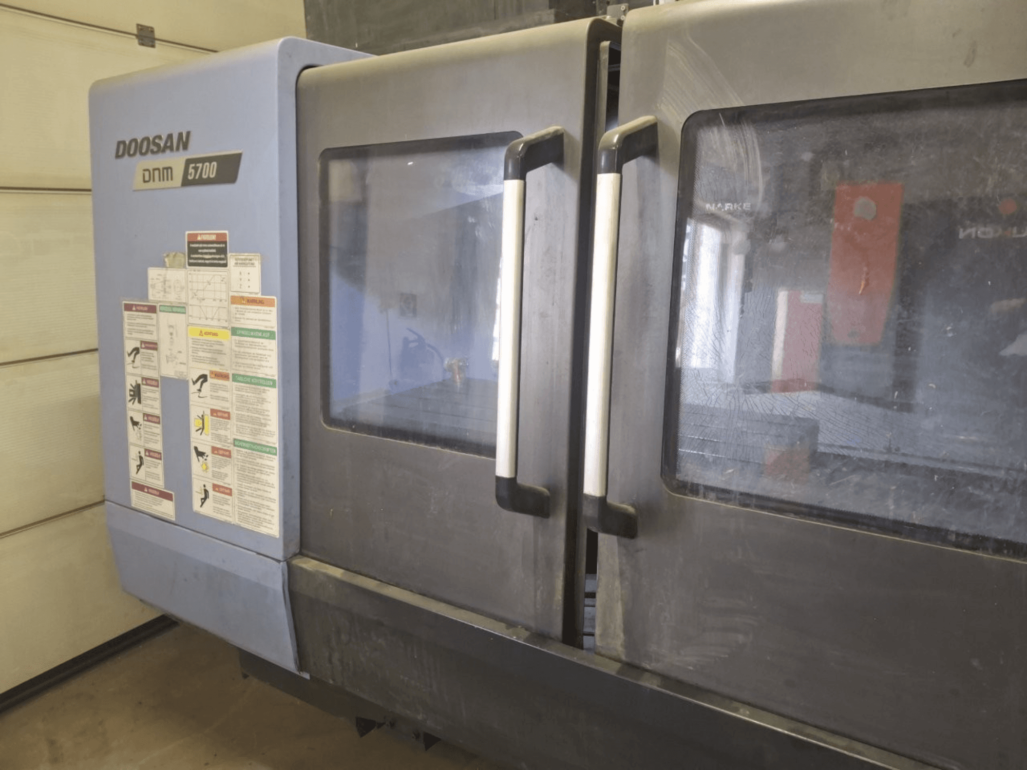Frontansicht eines DOOSAN DNM 5700 Vertikal-Bearbeitungszentrums mit Doppeltüren und Bedienfeldern mit Bedienhinweisen.