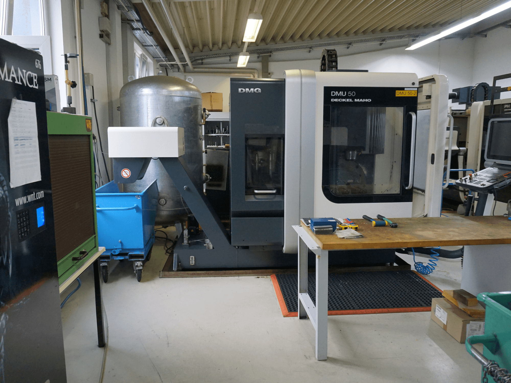 DMG DMU 50 Fräsmaschine in einer Werkstatt, Vorderansicht, mit Werkzeugen auf einem Holztisch und einer blauen Mülltonne auf Rädern in der Nähe.