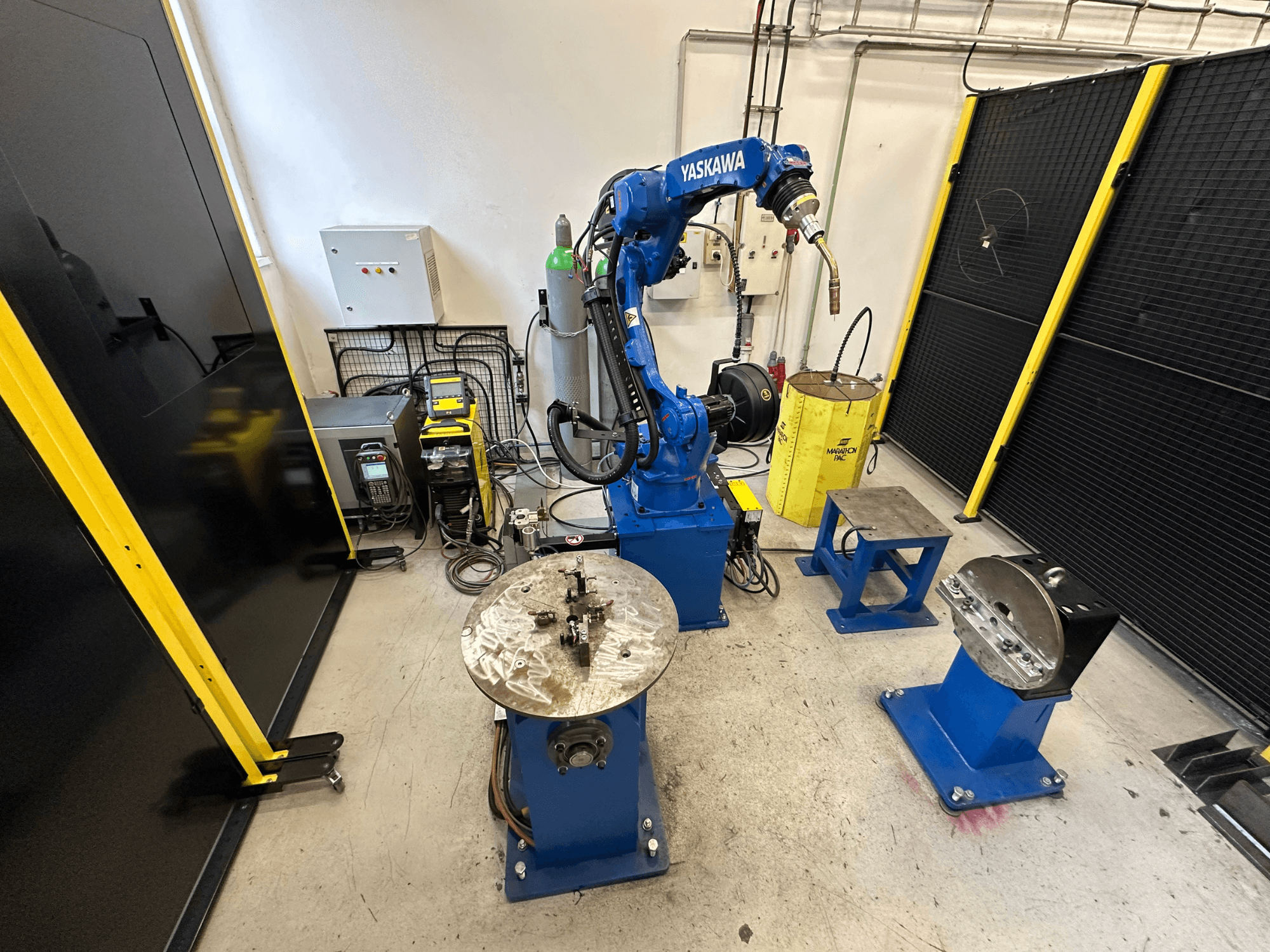 Blauer Yaskawa-Roboterarm mit zwei Arbeitstischen, Schweißgeräten und Sicherheitsbarrieren; Vorderansicht eines Industriearbeitsplatzes.
