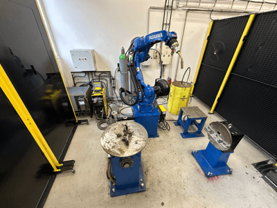 Blauer Yaskawa-Roboterarm mit zwei Arbeitstischen, Schweißgeräten und Sicherheitsbarrieren; Vorderansicht eines Industriearbeitsplatzes.