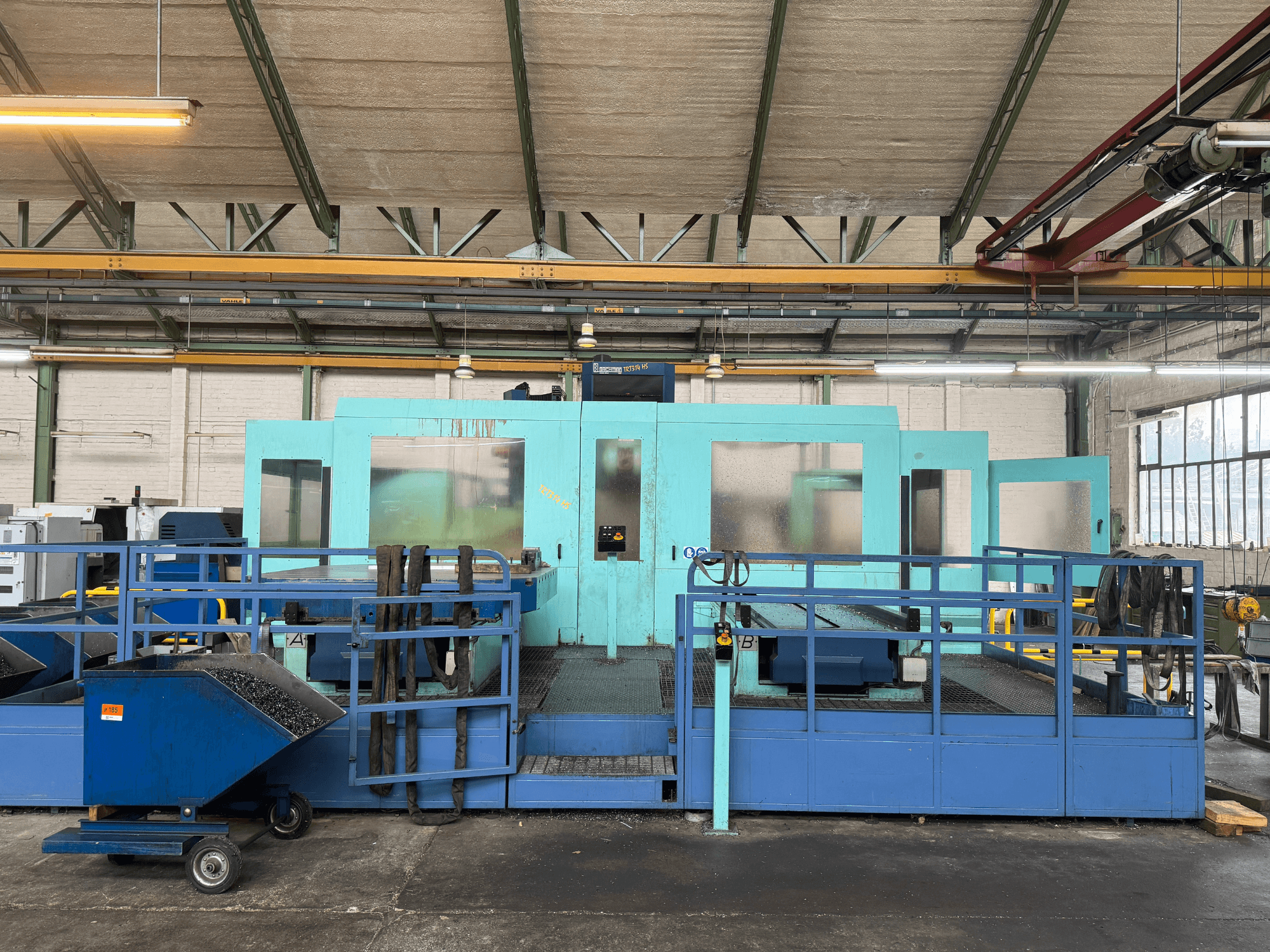 Blaue CNC-Industriemaschine mit Schutzplatten, Vorderansicht, mit Werkzeugablage und seitlichem Abfallsammelwagen.