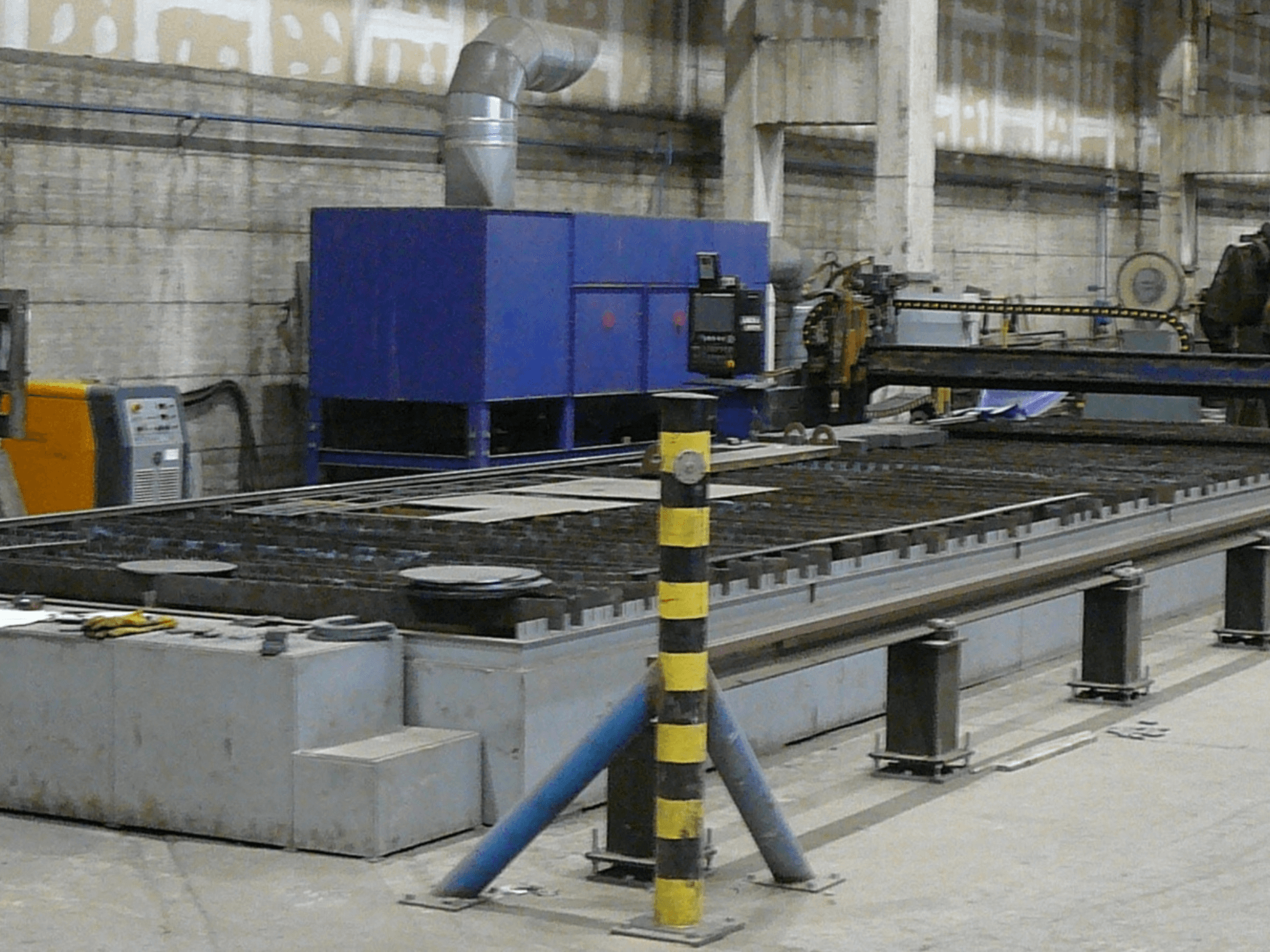 Einrichtung einer industriellen Metallschneidemaschine, Seitenansicht, mit blauem Schaltkasten, Sicherheitsstreifen und Metallarbeitsfläche.
