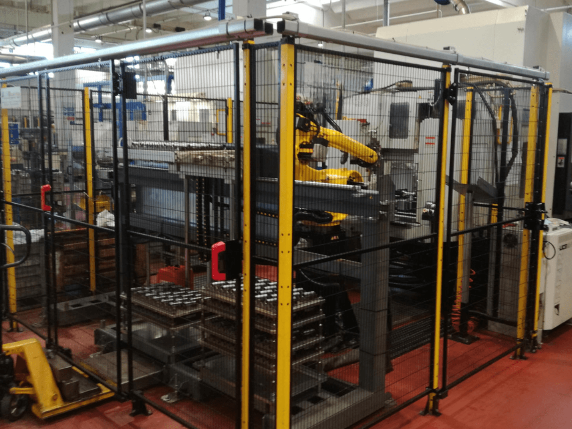 Vertikales Bearbeitungszentrum Enshu EV450 in einem umzäunten Arbeitsbereich, mit Roboterarmen und mehreren gestapelten Werkstücken. Vorderansicht.