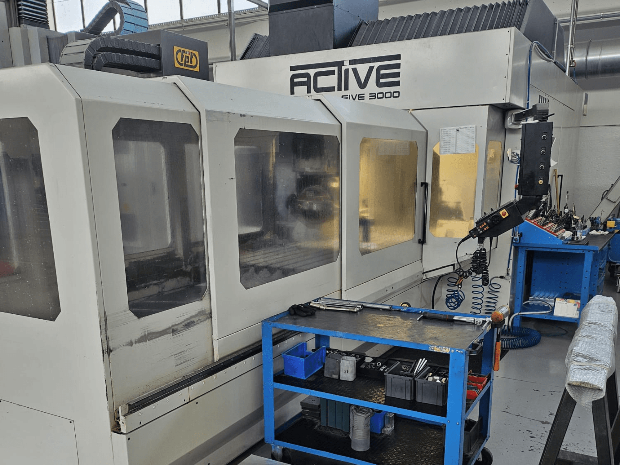 Aktive CNC-Maschine 3000 in einer Werkstatt, Vorderansicht mit Werkzeugen auf einem blauen Wagen, sichtbare Bedienelemente und Arbeitsbereich im Inneren.