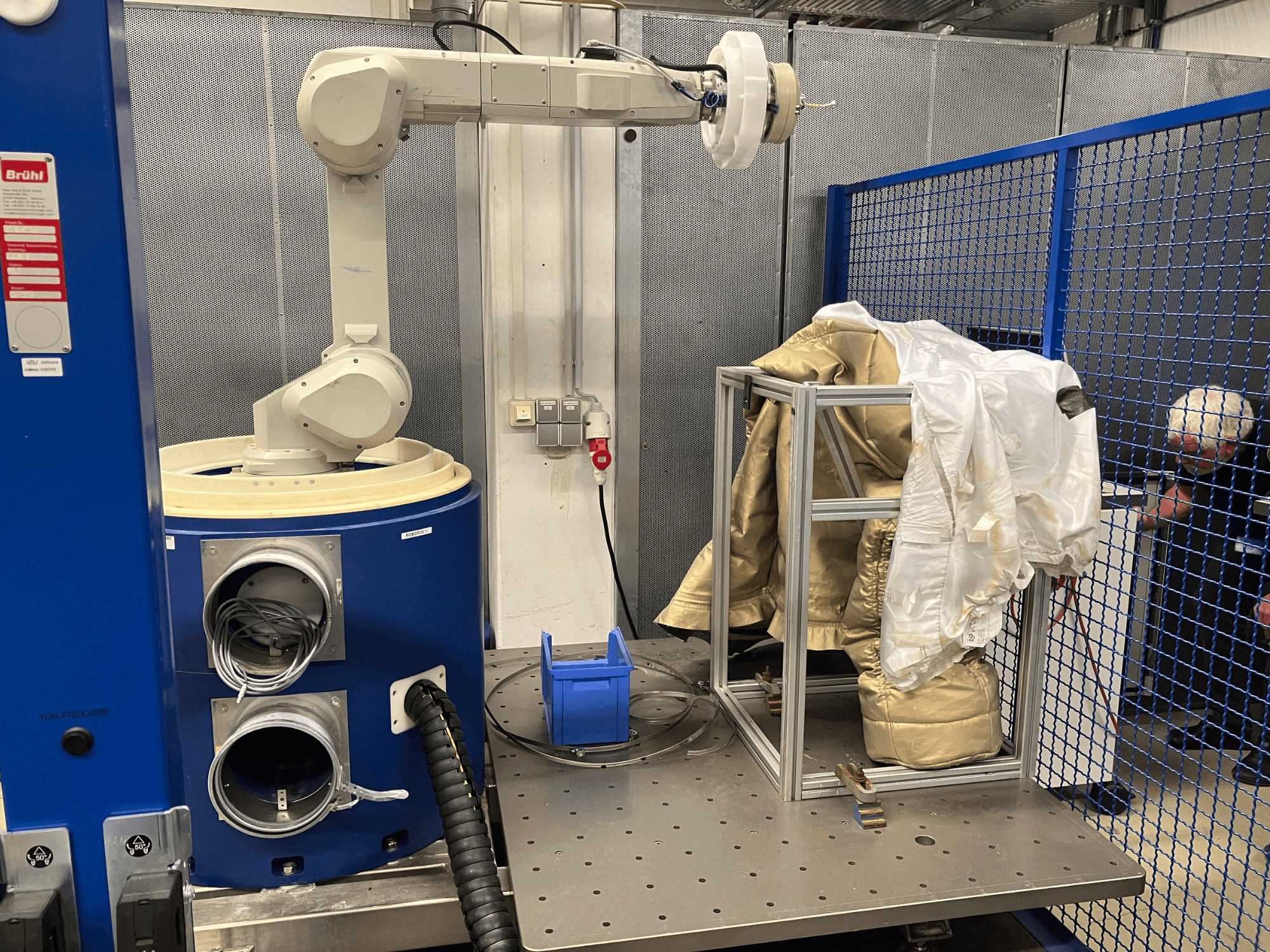Roboterarm in der Nähe einer blauen Kammer, der einen weiß-goldenen Anzug auf einem Metallrahmen hält, Seitenansicht mit Sicherheitsnetz im Hintergrund.
