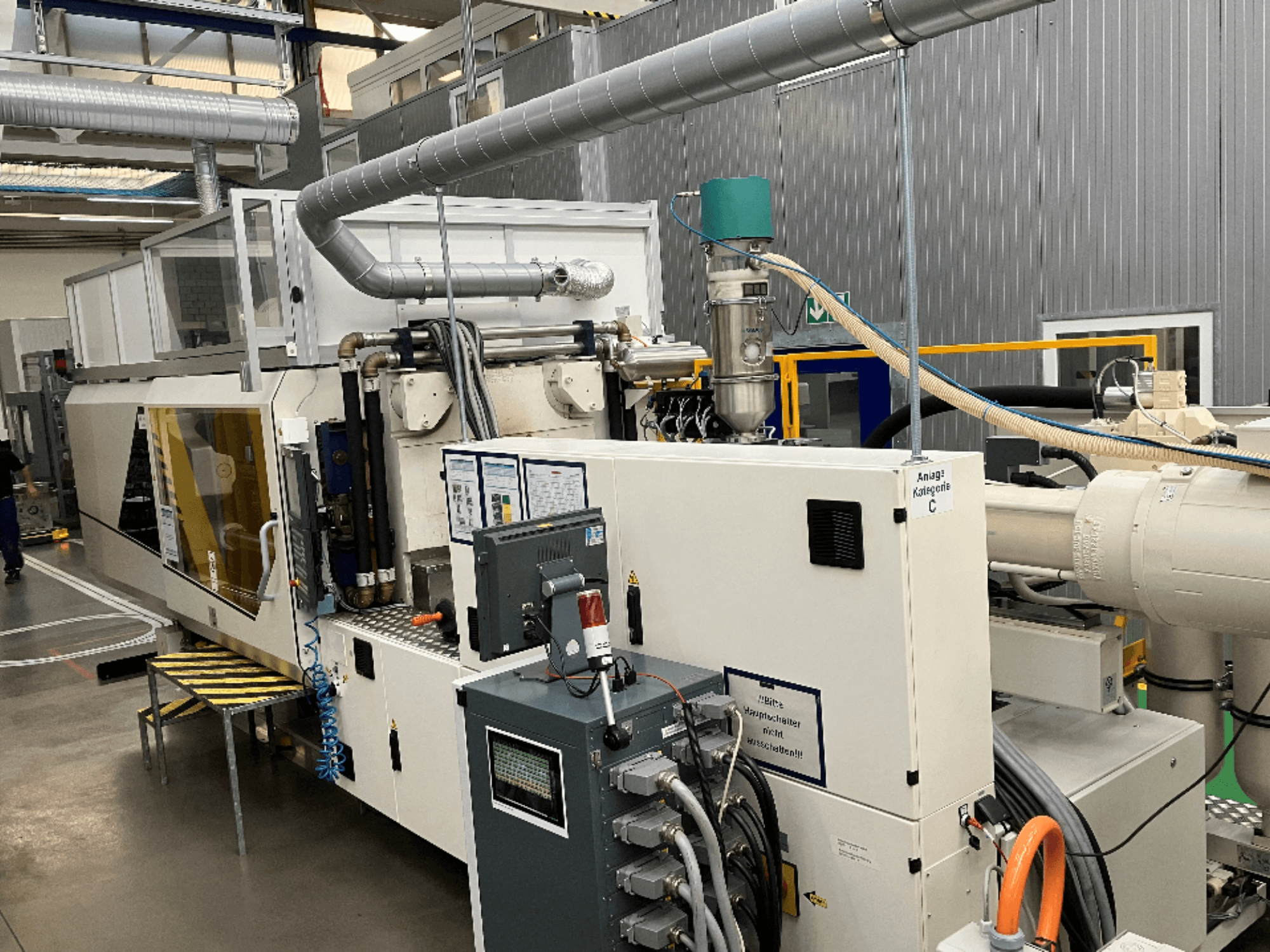 Spritzgießmaschine, Vorderansicht, mit Markenetikett, Bedienfeld und sichtbaren Schläuchen in einer Fabrikumgebung.