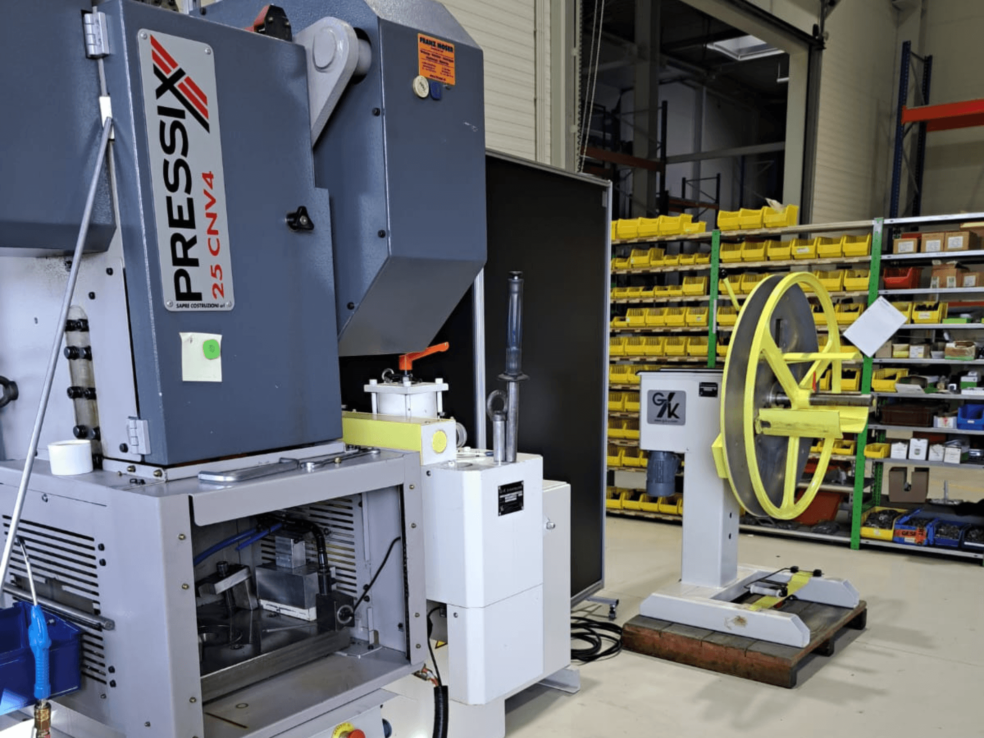 Industrieller Maschinenaufbau mit PRESSIX 25 CNV4 auf der linken Seite und einem gelben Rad auf einem grauen Sockel auf der rechten Seite, Vorderansicht.