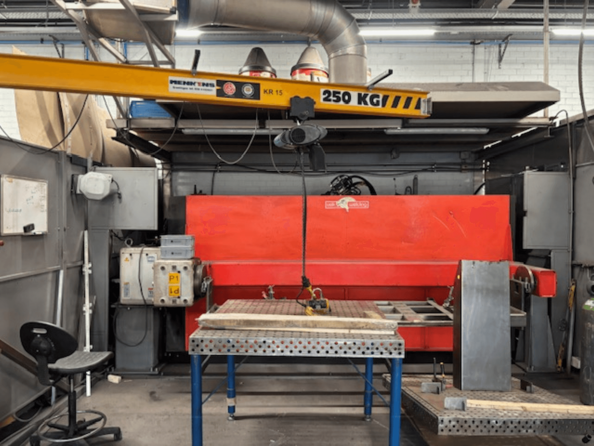 Industrielle Werkstatt mit einer roten Hydraulikpresse, einem Brückenkran mit der Aufschrift "KR 15" und einem Arbeitstisch aus Metall, Seitenansicht Perspektive.