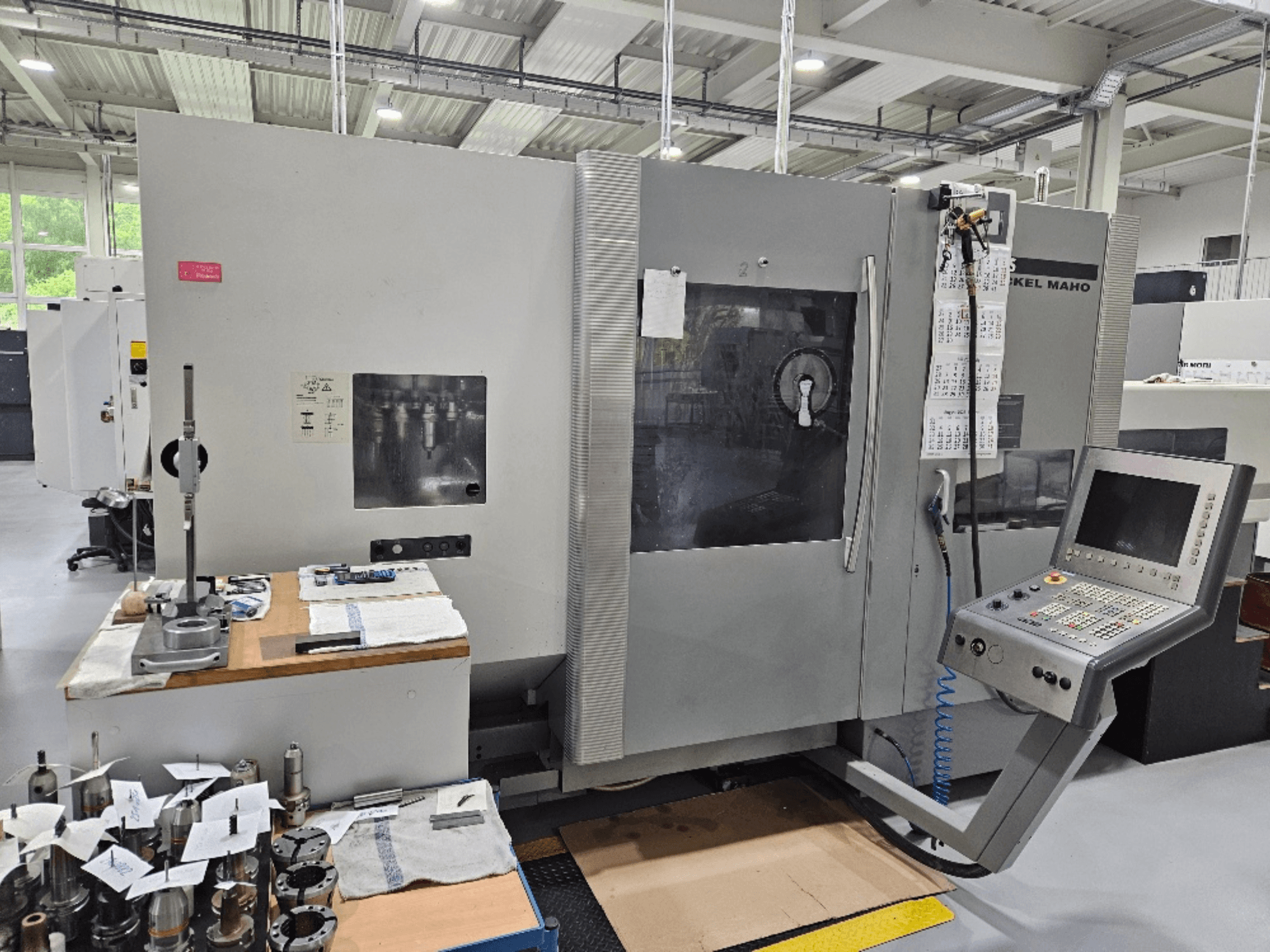 Frontansicht einer DMG Mori DMU 70 Maschine mit Bedienfeld, Werkzeugen auf einem Holztisch und verschiedenen Werkstücken in der Nähe.