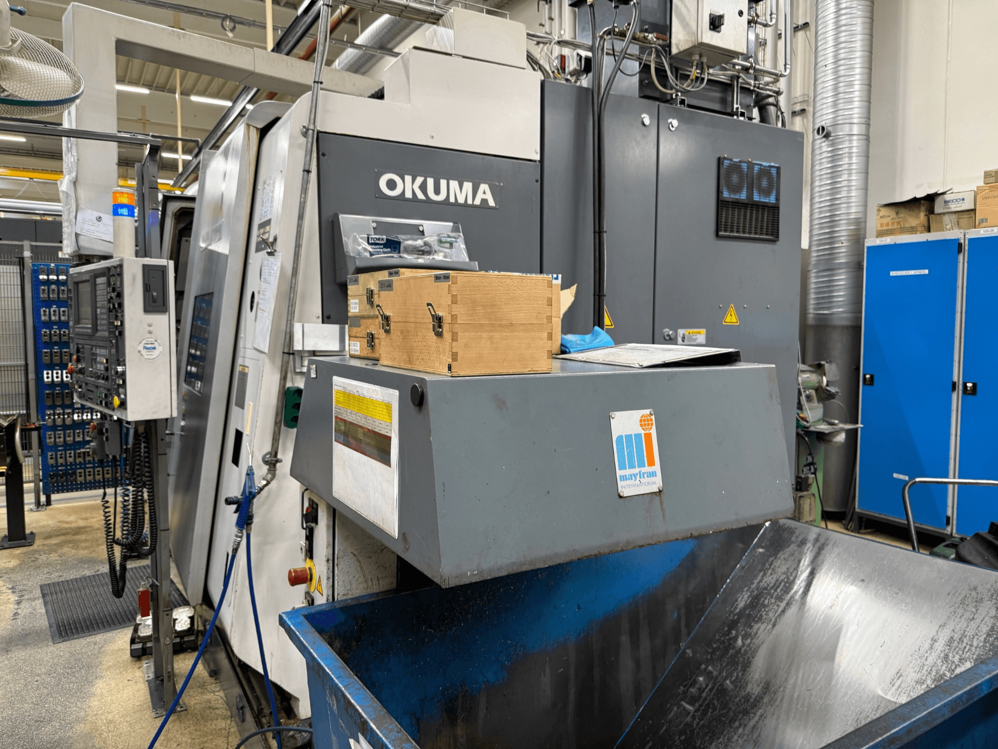 Okuma CNC-Maschine in der Fabrik, Vorderansicht; mit Bedienfeld, Holzkästen und blauem Abfallbehälter mit Mayfran-Logo.