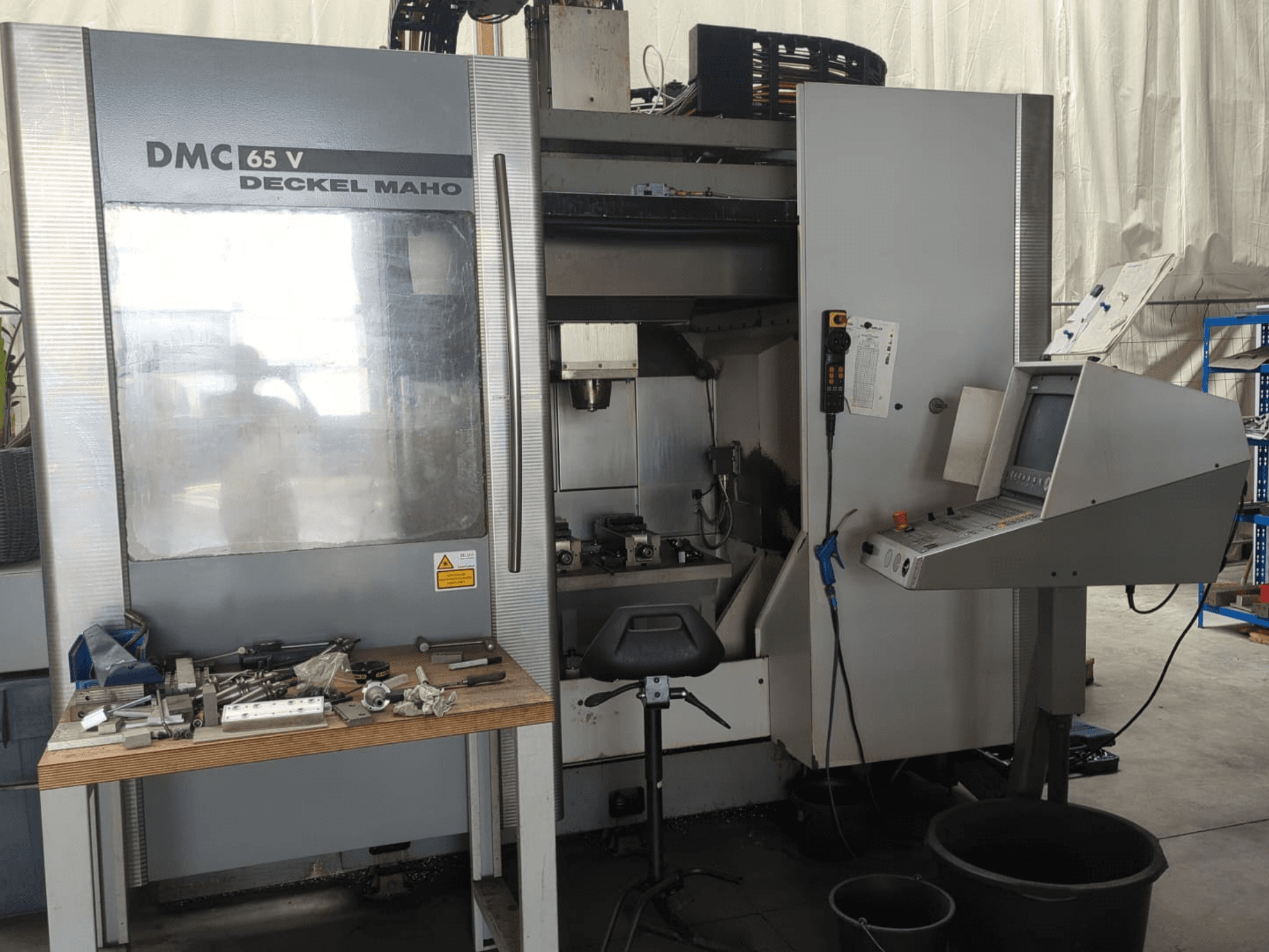 Vorderansicht eines vertikalen Bearbeitungszentrums vom Typ DECKEL MAHO DMC 65 V mit Bedienfeld, Werkzeugen auf einem Tisch und eingerichteter Arbeitsfläche.