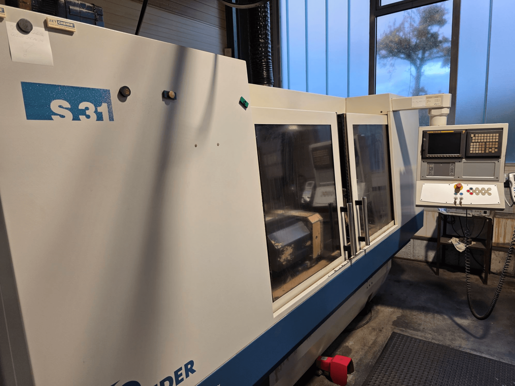 S31 CNC-Schleifmaschine, Vorderansicht, mit Bedienfeld mit Bildschirm und Tasten, große Glastür, Industrieumgebung.