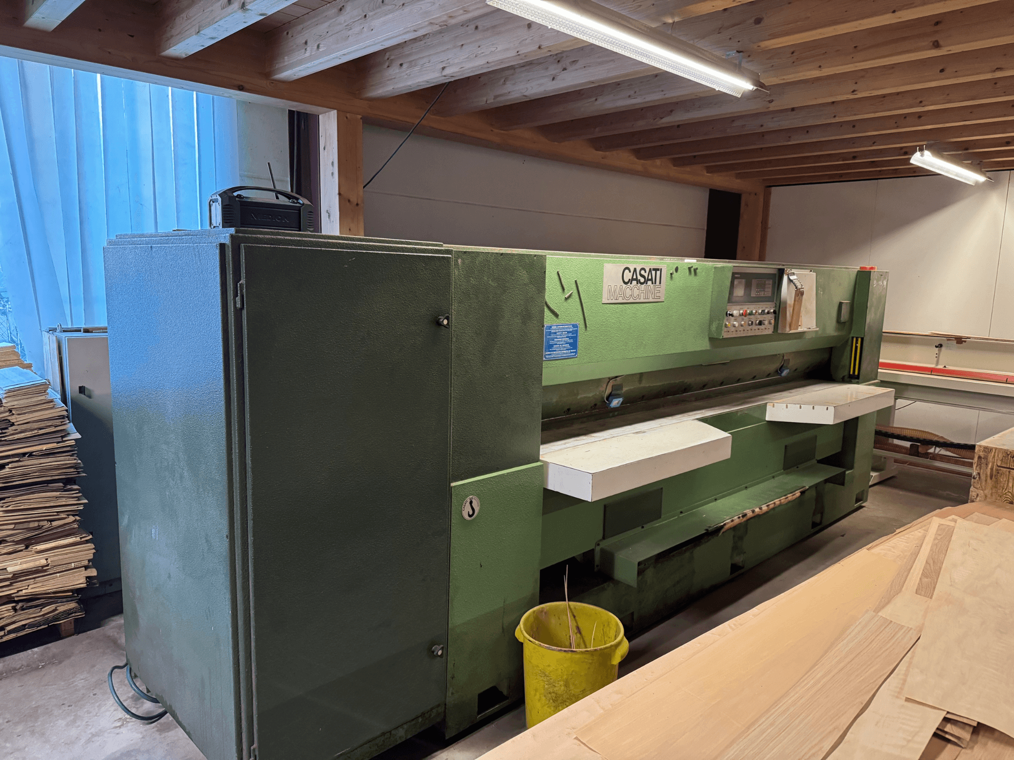 Grüne CASATI Holzbearbeitungsmaschine in einer Werkstatt, Vorderansicht, mit Bedienfeld und weißen Arbeitsflächen, umgeben von Holzstapeln.