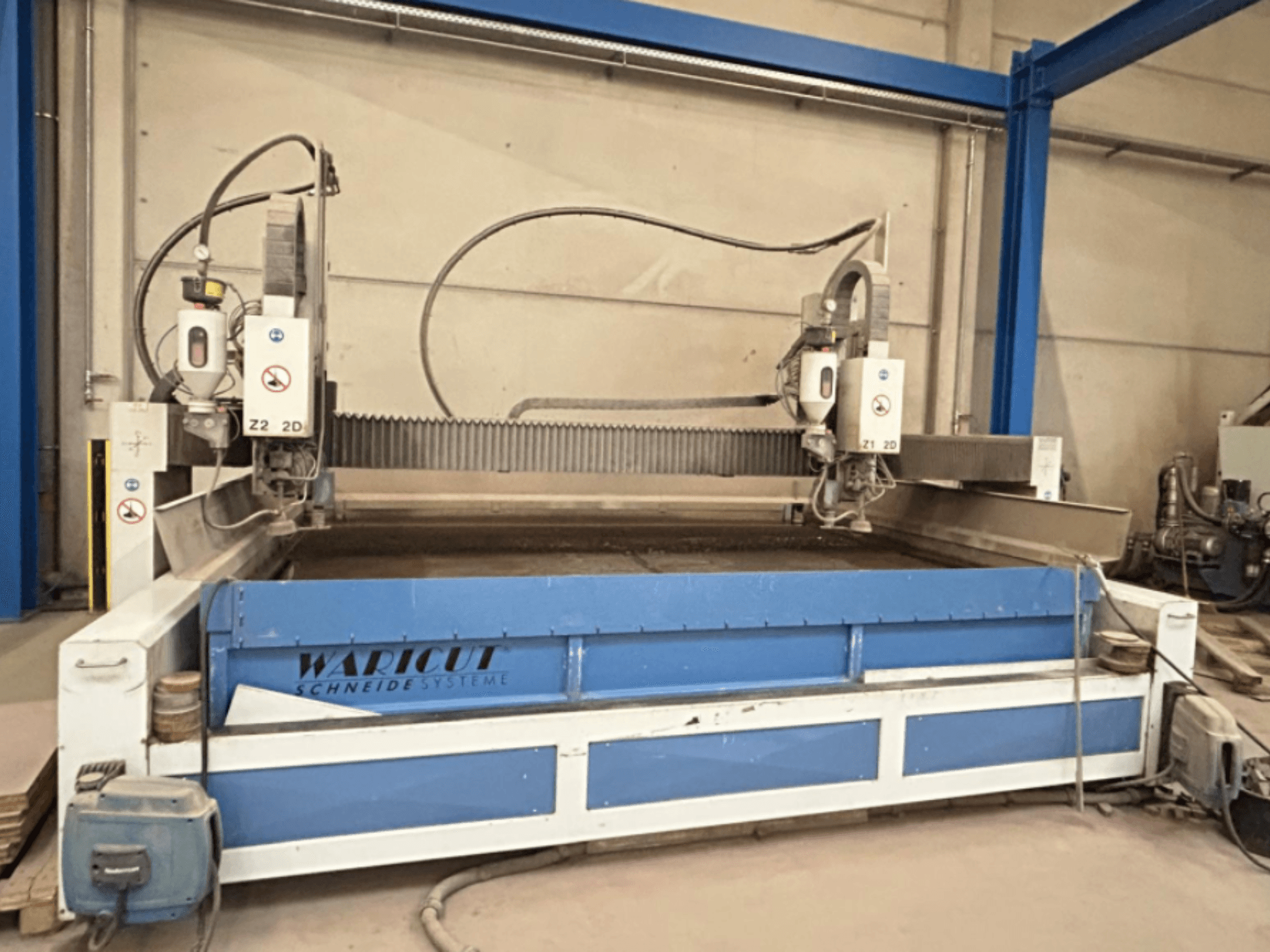 Wasserstrahlschneidmaschine von Waricut, Vorderansicht; mit zwei Schneidköpfen und blauer Arbeitsfläche in einer Werkstatt.