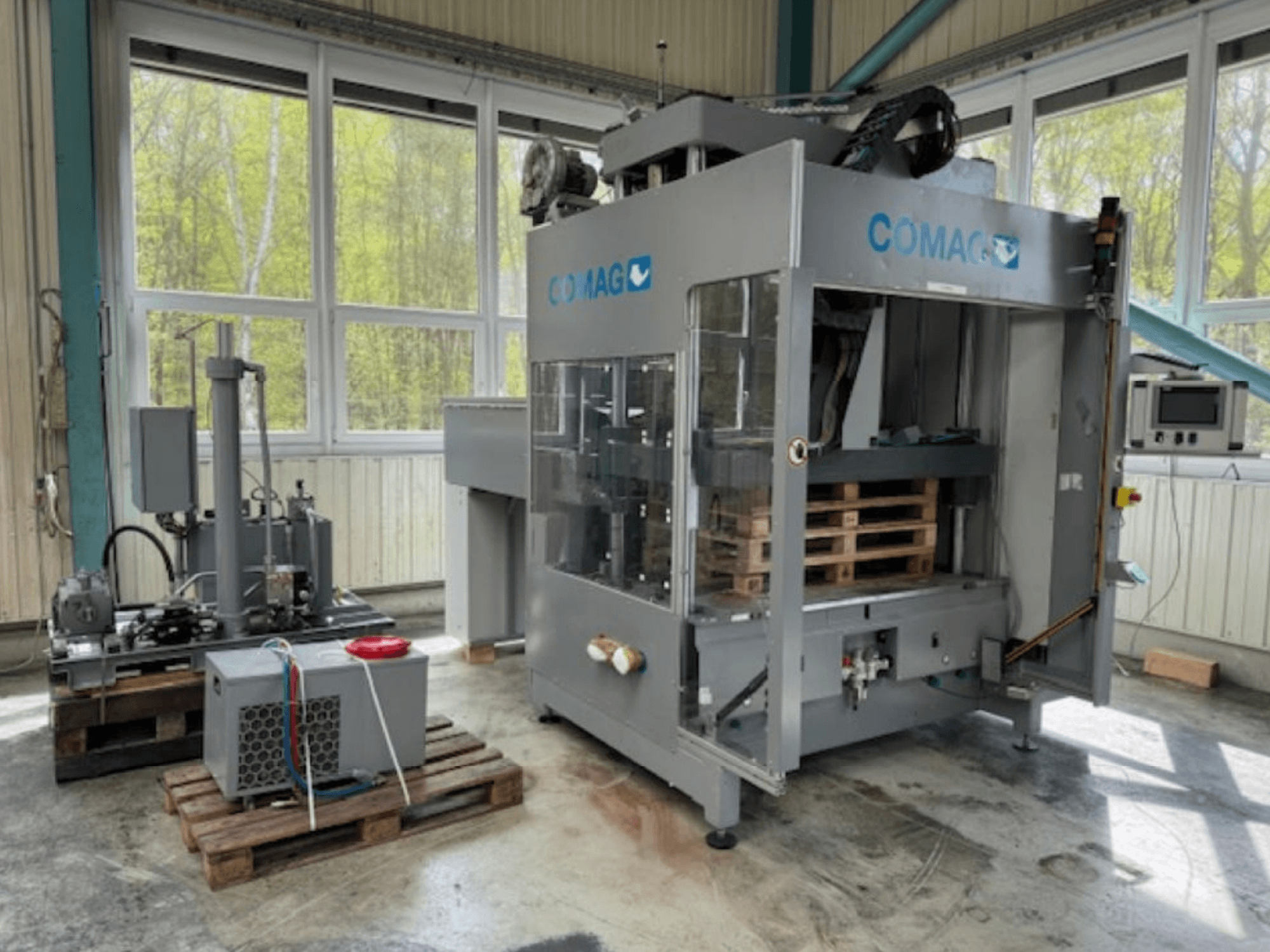 COMAG Palettenwickelmaschine in Industrieumgebung, Vorderansicht; Paletten und hydraulische Komponenten sichtbar.