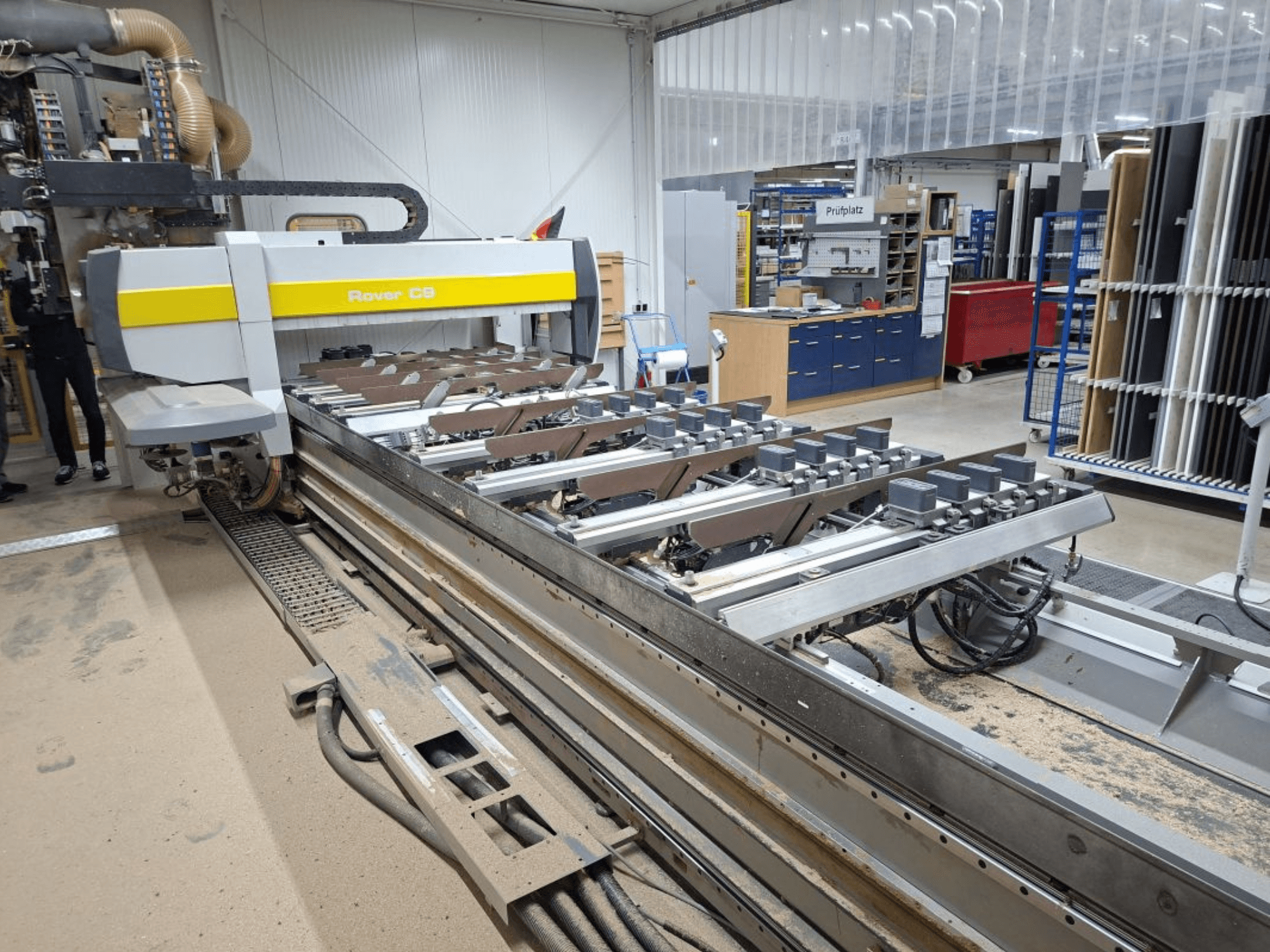 Rover C9 CNC-Maschine in einer Werkstatt; Vorderansicht zeigt Schneidwerkzeuge und Fördersystem, mit Staubansammlung auf dem Boden.