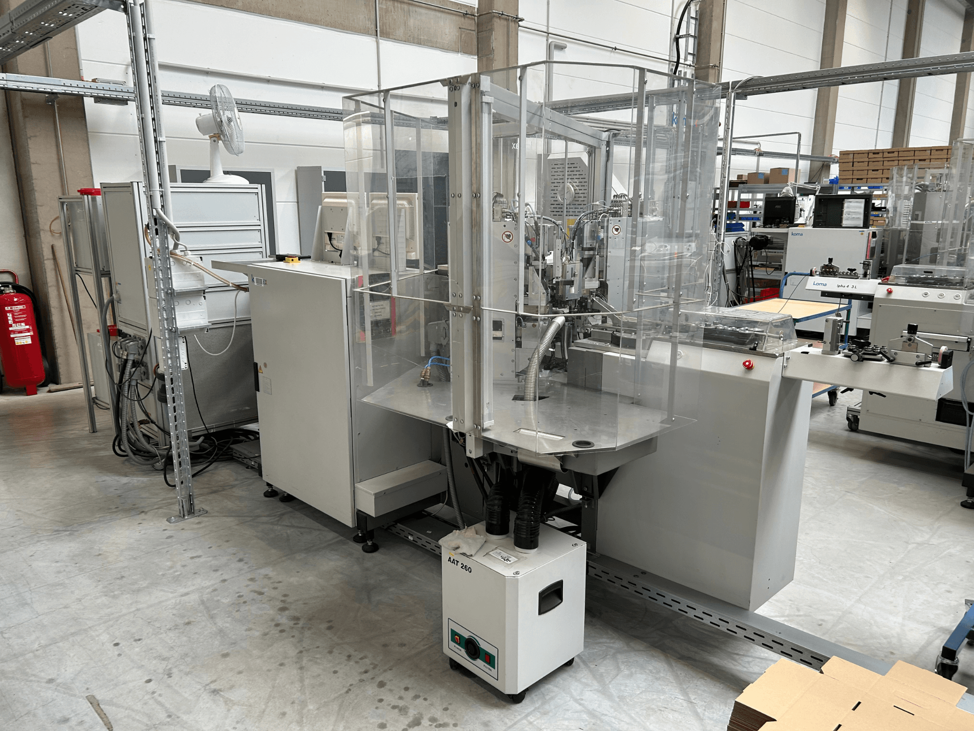 KOMAX Alpha 433 L-Automatisierungsanlage in einer Fabrikumgebung. Mit transparenter Sicherheitsabdeckung und verschiedenen Bedienfeldern.