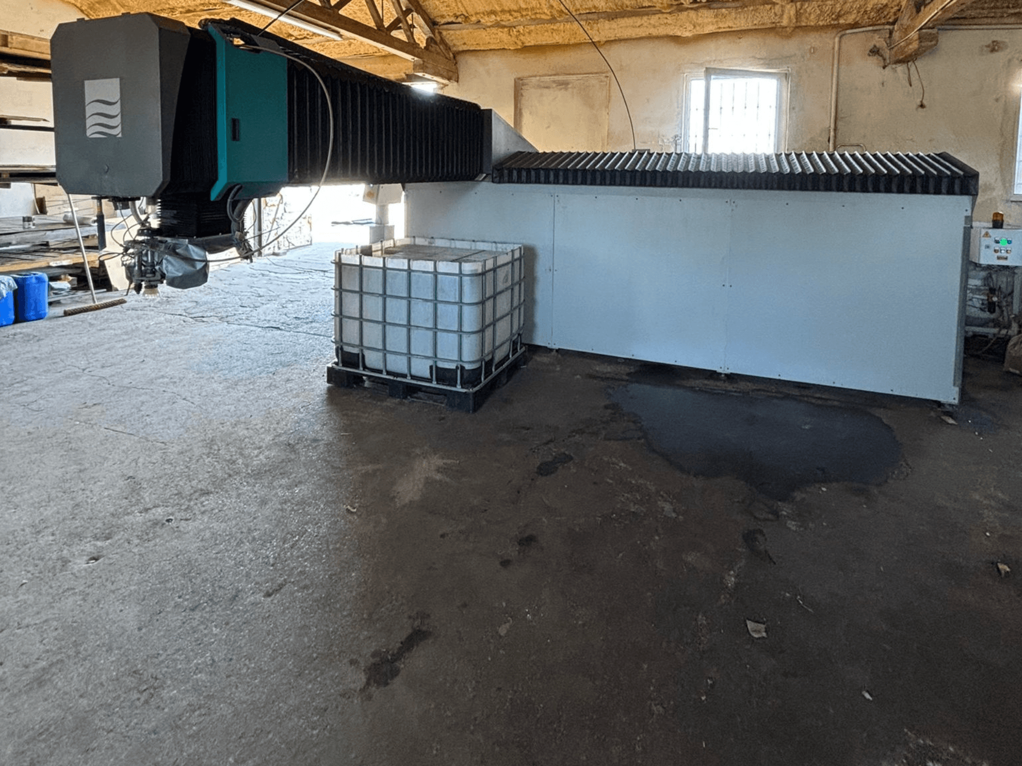 Industrielle Wasserstrahlmaschine in einer Werkstatt, Seitenansicht, mit einem großen Wassertank und einem Bedienfeld daneben.