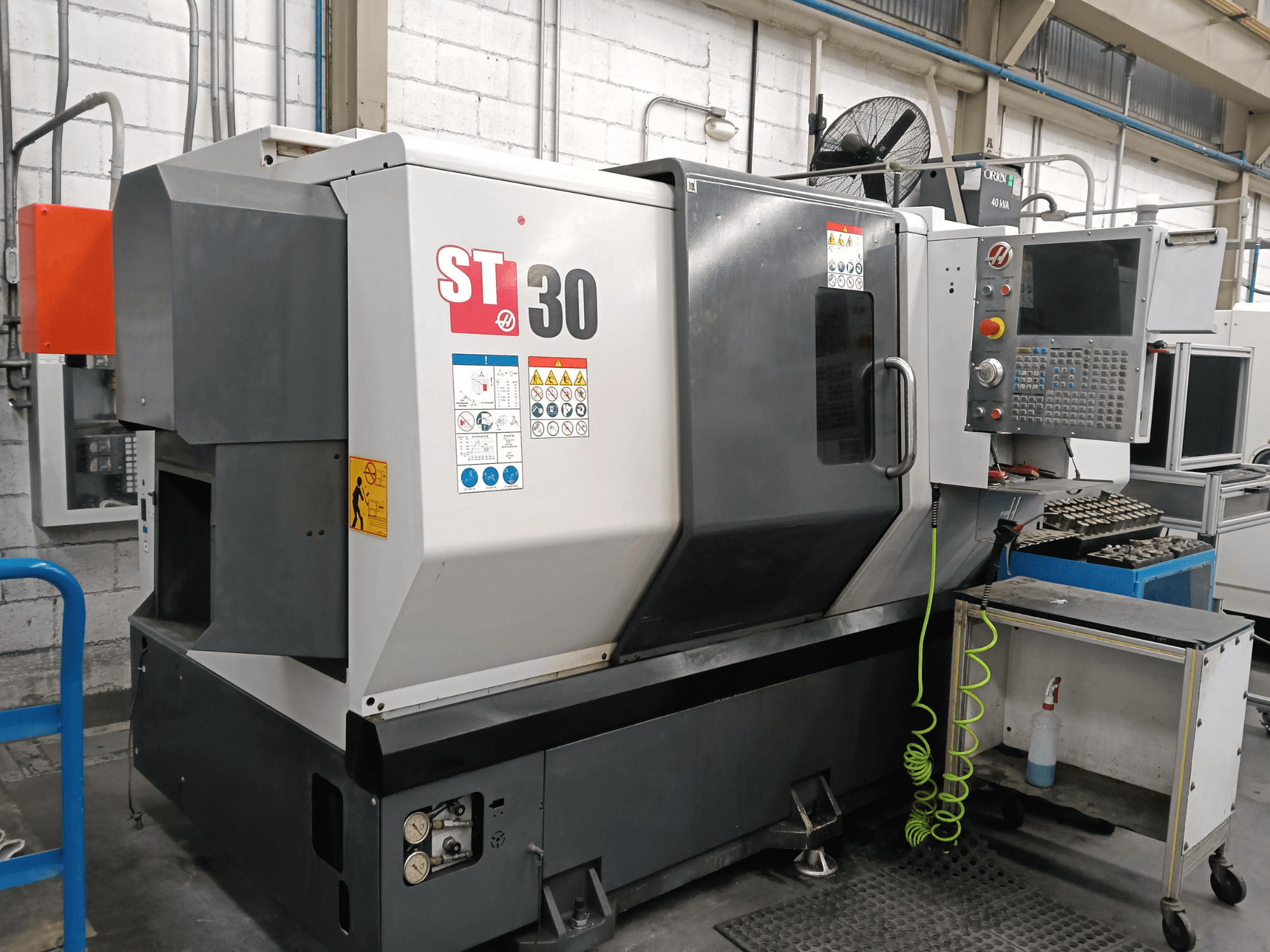 Alternativtext: Vorderansicht der horizontalen Drehmaschine HAAS ST-30 mit Bedienfeld, Sicherheitsaufklebern und Werkzeugen auf einem daneben stehenden Wagen.