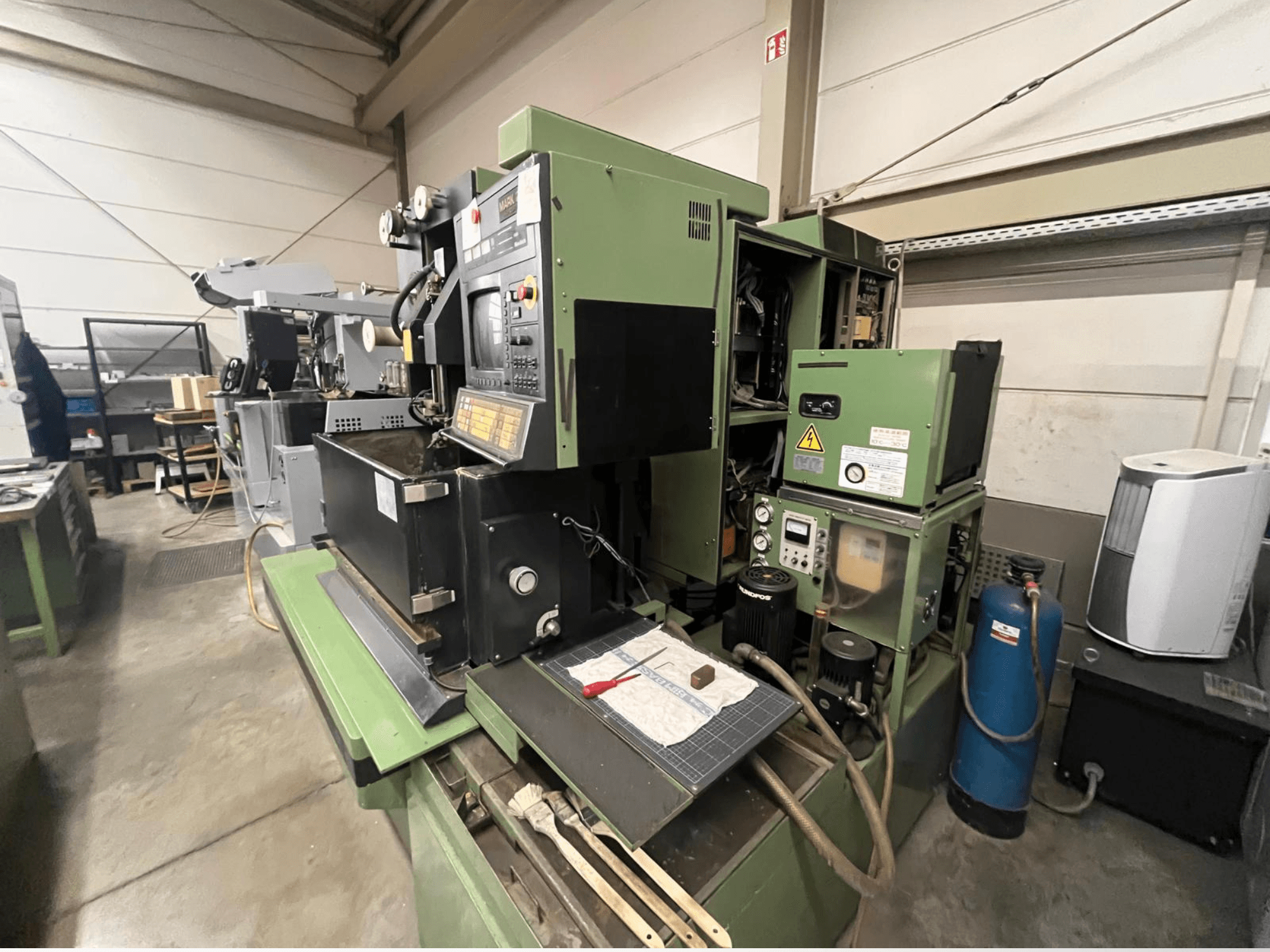 Grüne Industriemaschine mit Bedienpult, Seitenansicht; einschließlich Hydraulikkomponenten und Arbeitsfläche mit Werkzeugen.