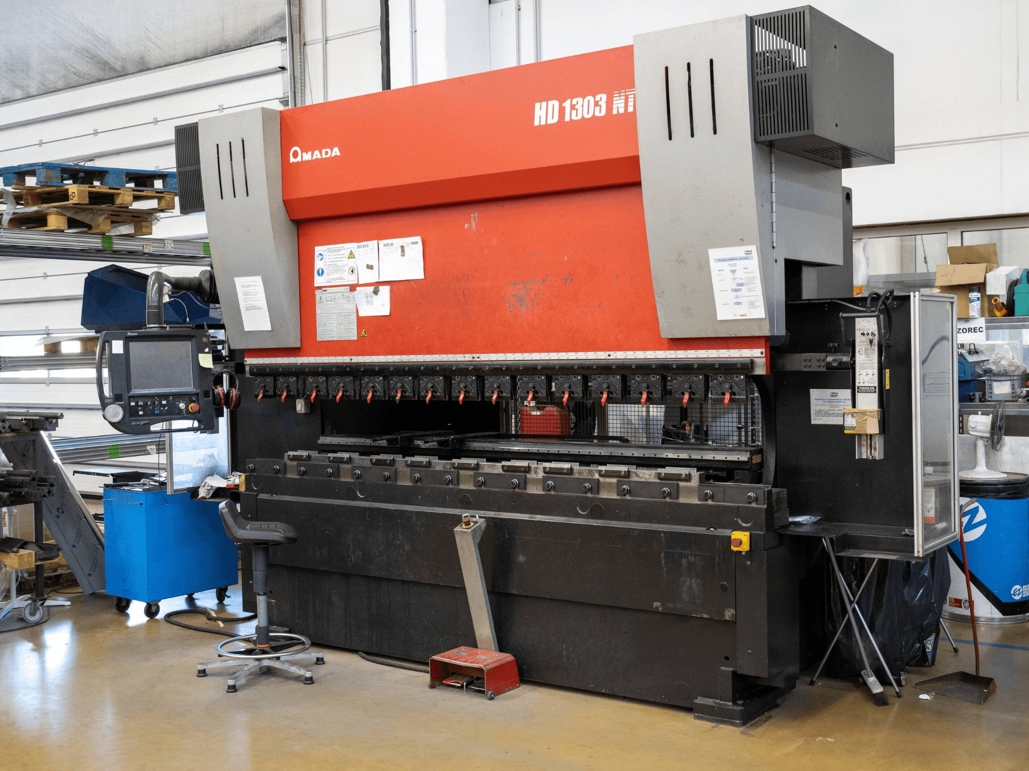 Hydraulische Abkantpresse Amada HD 1303 in einer Werkstatt, Vorderansicht; mit mehreren Spannvorrichtungen, Bedienfeld und verstellbarem Hocker.