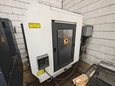 Industrielle CNC-Maschine, Seitenansicht; mit Bedienfeld, Warnschildern und Holzteilen auf einer Arbeitsfläche.